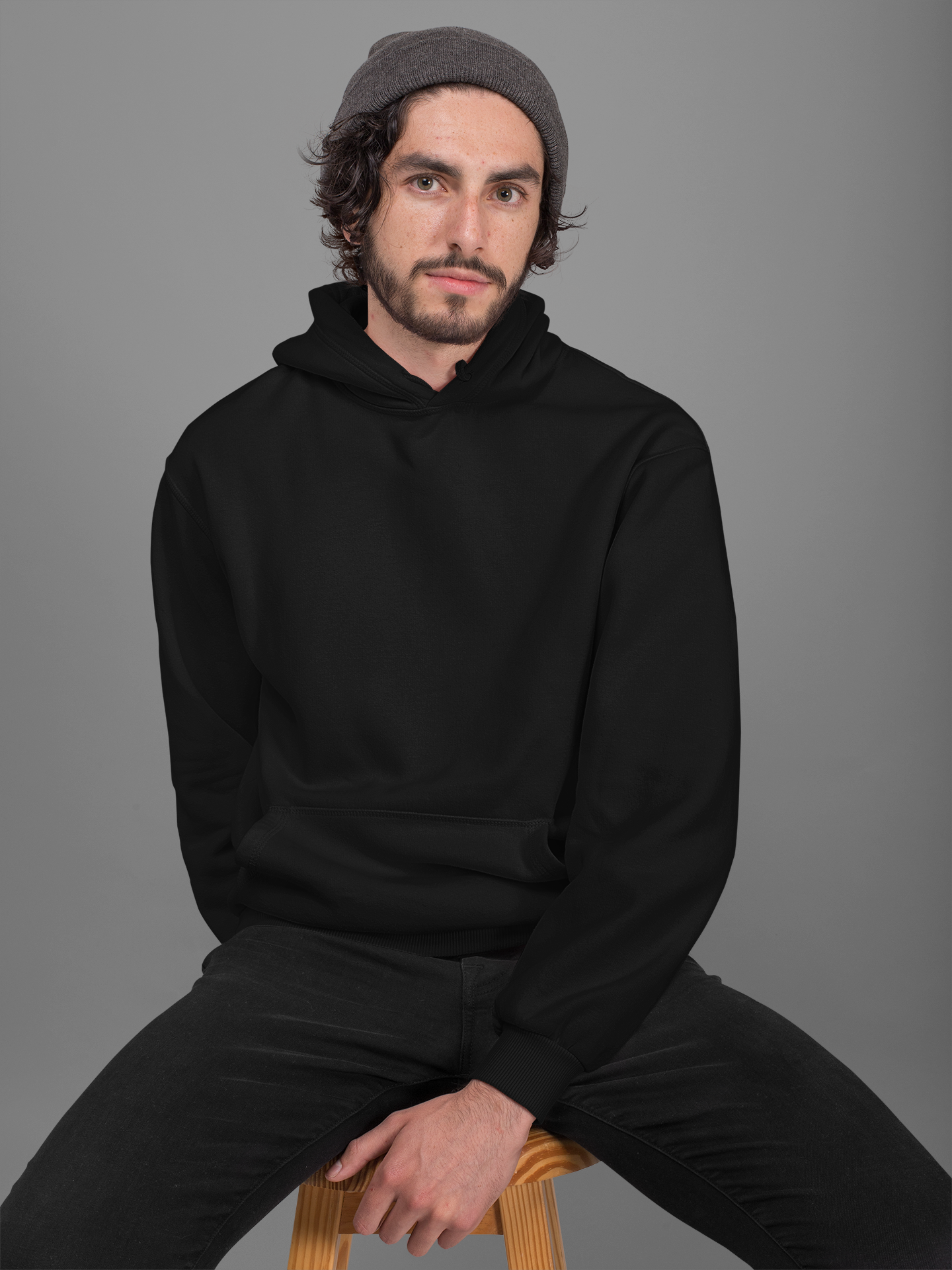A male model wearing a black hoodie with a hood, sitting on a chair.