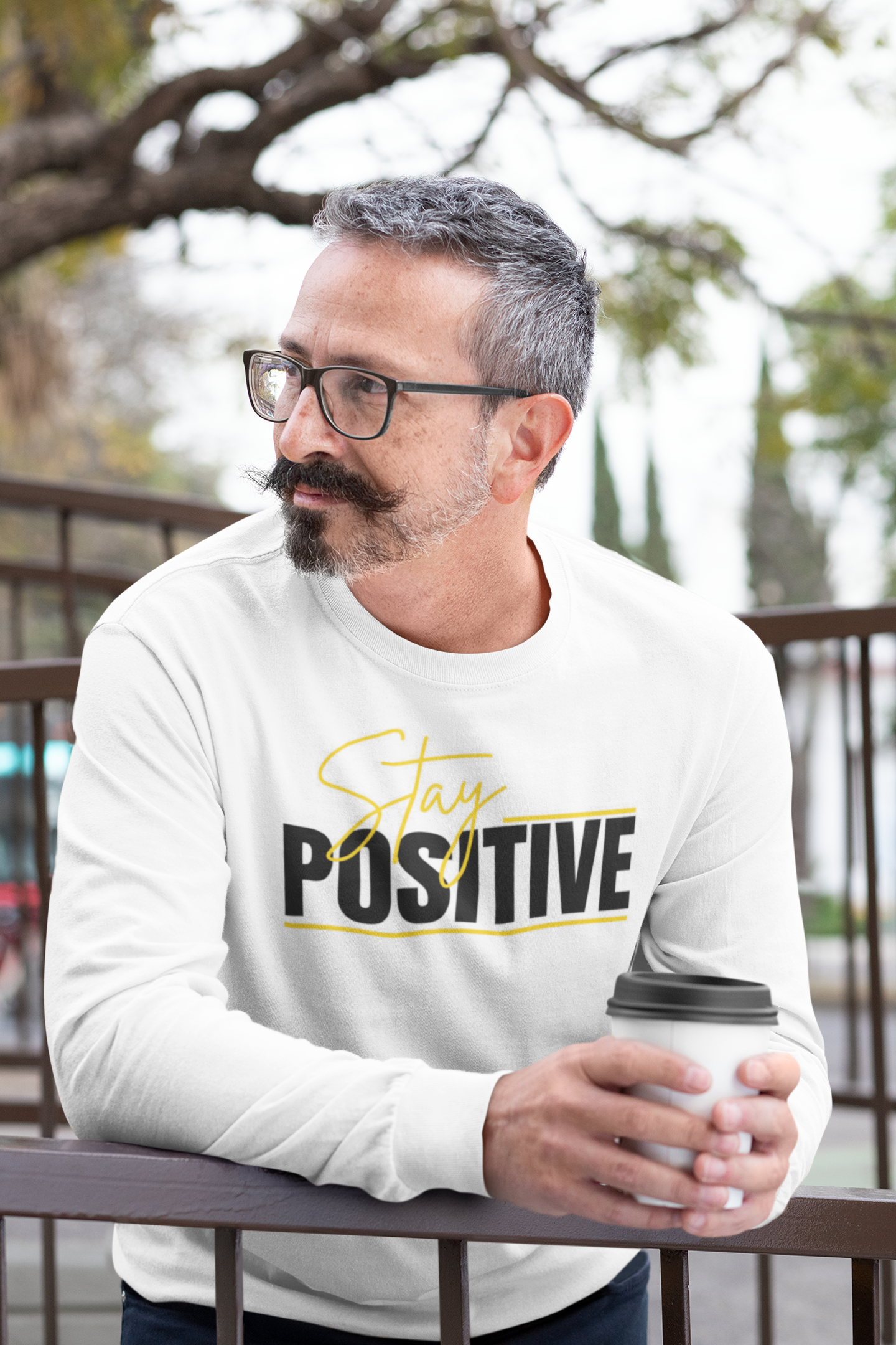 Man wearing a white sweatshirt with 'Stay Positive' text, holding a coffee cup outdoors.
