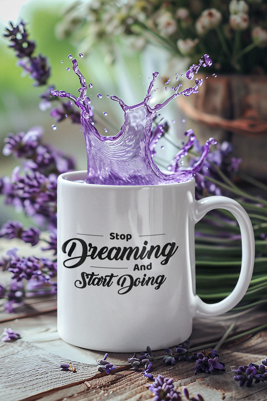 A white ceramic mug with the words 'Stop Dreaming Start Doing' printed in black. The mug is accompanied by a splash of purple liquid, and there are lavender flowers around it.