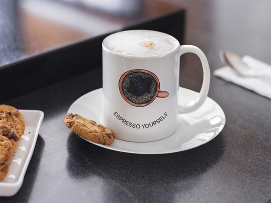 A white ceramic mug with 'ESPRESSO YOURSELF' text and a coffee bean graphic on the front, accompanied by a saucer and a cookie, placed on a black surface.
