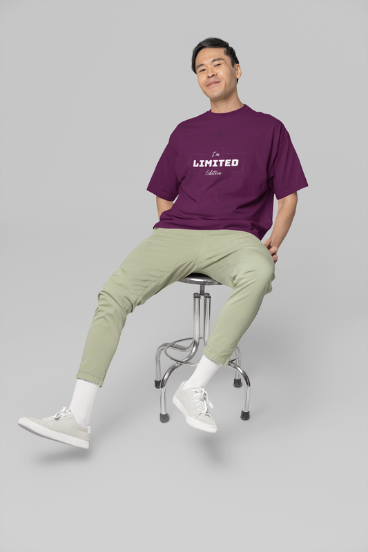 Man sitting on a stool wearing a purple t-shirt and light green pants against a gray background