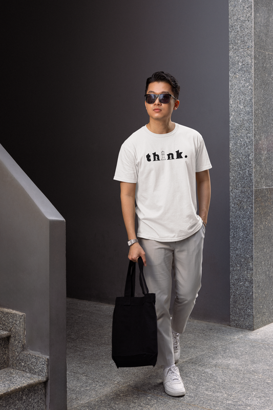 Man wearing a white t-shirt with 'think.' text, holding a black bag, standing against a gray wall.